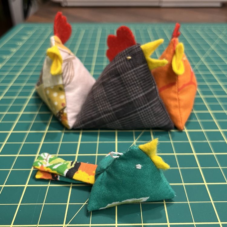Photo of the same 3 chickens in the background, but there's a smaller fabric bird with tail in the foreground. The little bird is mostly kelly green with a multi-coloured tailed with orange/white/yellow. They have a yellow beak made from felt, white embroidered eyes, and a little loop on the top of their head to use like a charm/keychain.
