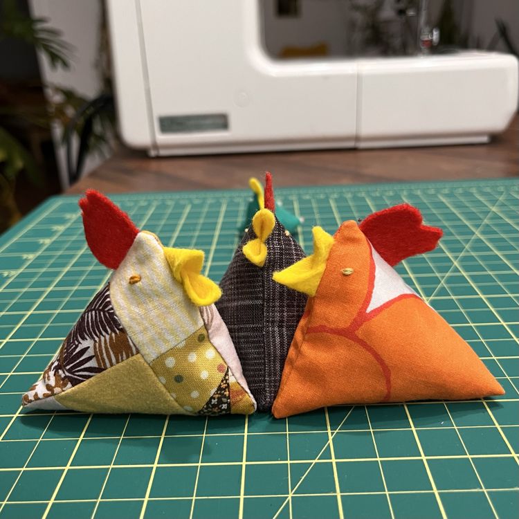 photo of 3 fabric chickens that are shaped like a triangle. One is beige/yellow/brown patchwork, another is dark brown/blue/white yarn-dyed plaid, and the last is primarily orange with a bit of white. They all have red combs and yellow beaks made from felt and embroidered yellow eyes.